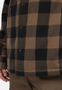 Brun och svart rutigt flanellskjorta med knappstängning, en texturerad tygkänsla och rak fåll. Närbild av mönstret.