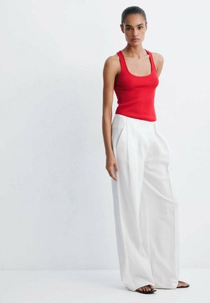 Femme portant un haut rouge sans manches, un pantalon blanc large taille haute et des sandales noires, debout devant un fond clair uni.