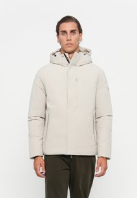 Giacca trapuntata di un beige chiaro con cappuccio, chiusura frontale con zip, tasche laterali e design minimalista. Tessuto liscio e forma ben definita.
