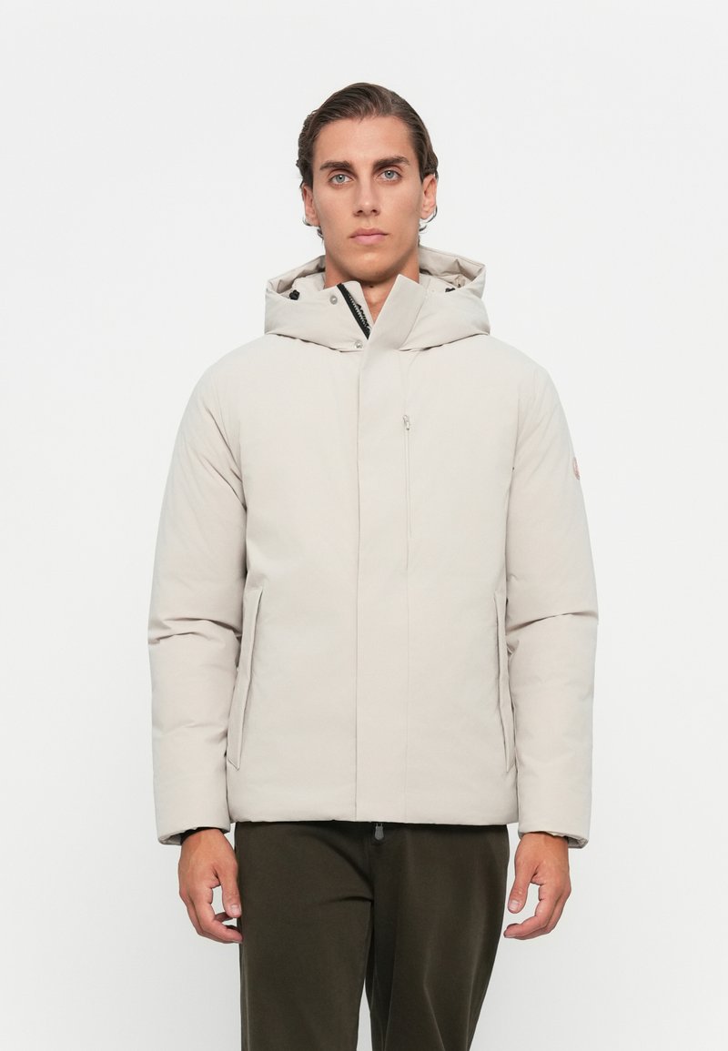 Giacca trapuntata di un beige chiaro con cappuccio, chiusura frontale con zip, tasche laterali e design minimalista. Tessuto liscio e forma ben definita.