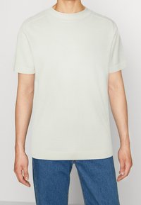 Světle béžové tričko s krátkým rukávem vyrobené z měkké tkaniny, s kulatým výstřihem, bezšvovým designem ramen a pohodlným střihem. Kombinováno s modrými džíny.