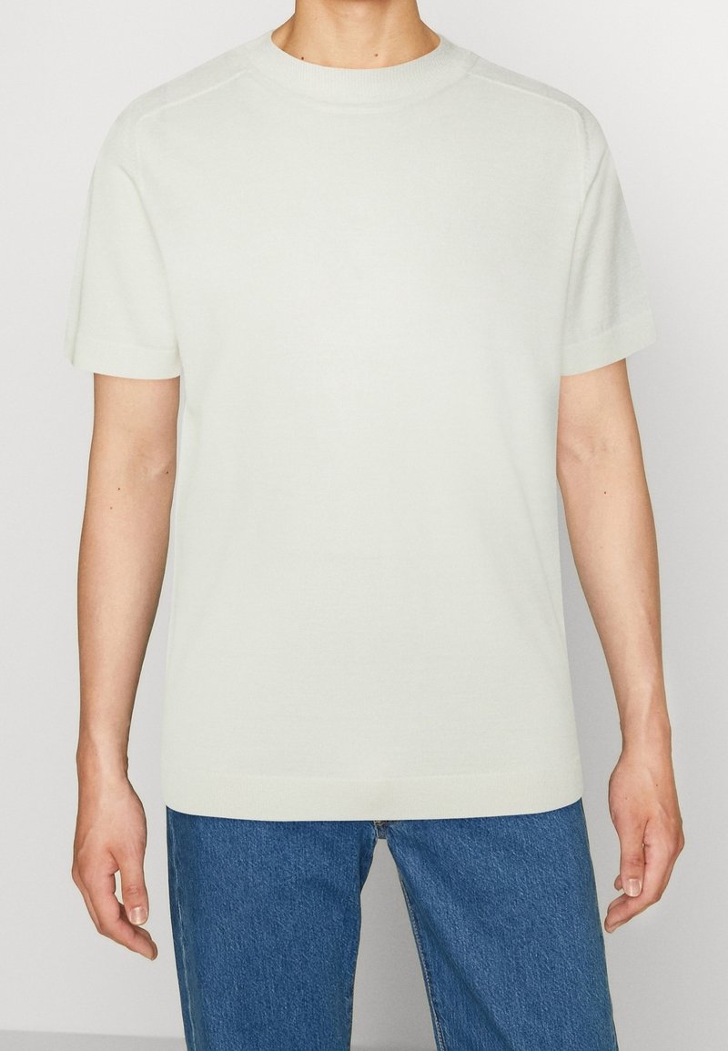 Světle béžové tričko s krátkým rukávem vyrobené z měkké tkaniny, s kulatým výstřihem, bezšvovým designem ramen a pohodlným střihem. Kombinováno s modrými džíny.