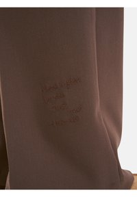 Brauner Stoff mit einer glatten Textur zeigt prägierte Schriftzüge: "Habe einen Plan. Vertrauens du mir? -Universum." Schlichtes Design, ohne zusätzliche Akzente.
