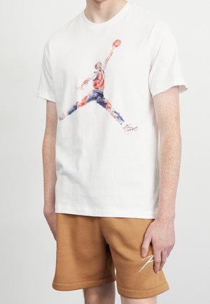 T-shirt blanc avec graphisme d'un joueur de basketball en pleine suspension dunkant, style aquarelle, porté avec un short marron clair.