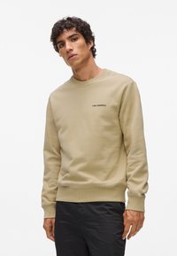 Béžová mikina s okrúhlym výstrihom, rebrovanými manžetami a priliehavým dolným lemom. Na ľavej strane hrude má čierny text loga. Hladká textúra.