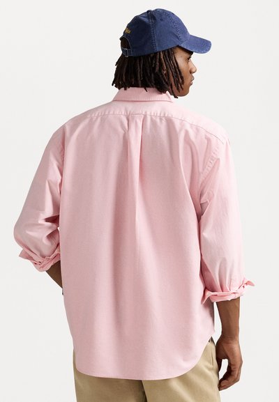 Chemise boutonnée rose clair avec des manches retroussées, présentant un pli au dos et un ourlet arrondi, associée à une casquette bleu foncé.