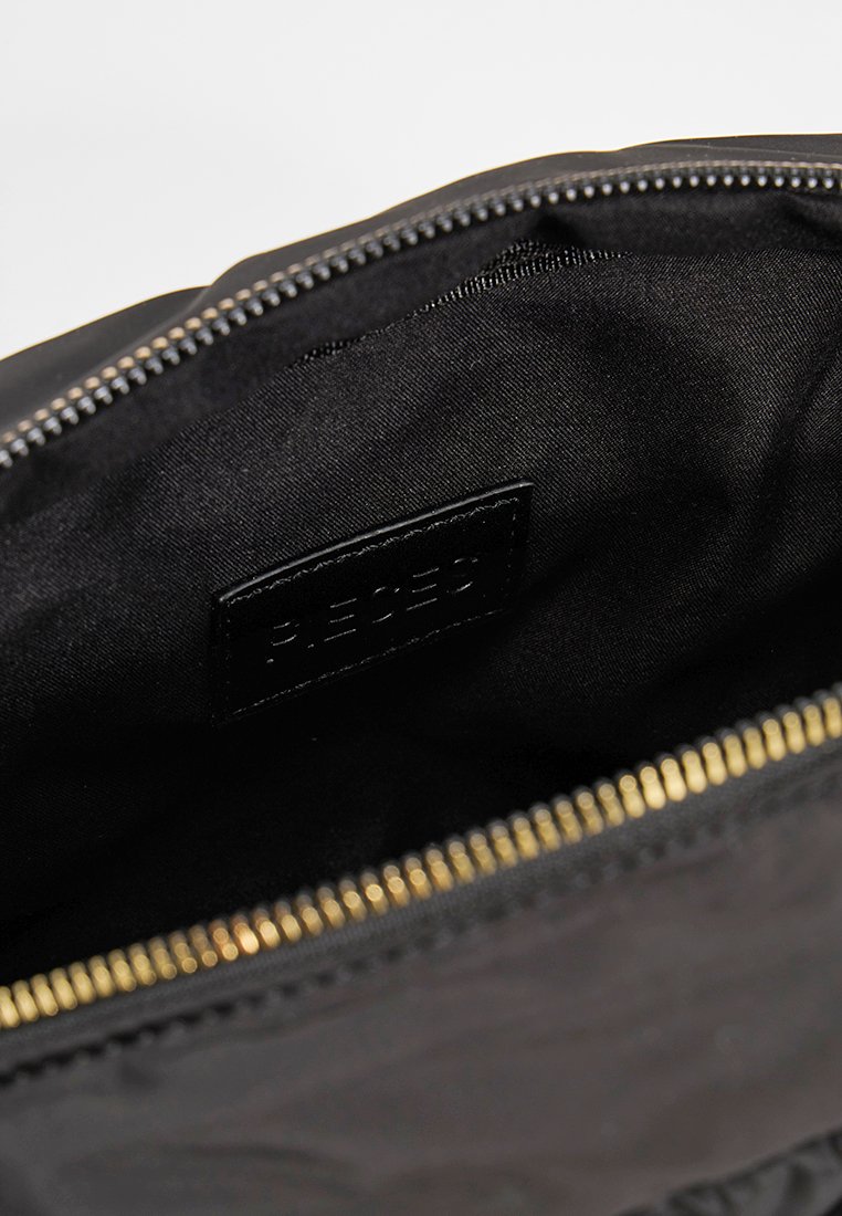 Wnętrze czarnej torby materiałowej o gładkiej fakturze, złoty zamek błyskawiczny oraz mała, tłoczona czarna metka. Podszewka jest również czarna i matowa.