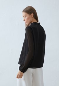 Zwarte blouse met lange mouwen en een geplooide halslijn, gemaakt van doorzichtige stof. Bevat strakke manchetten en heeft een relaxte pasvorm, gedragen met witte broeken.