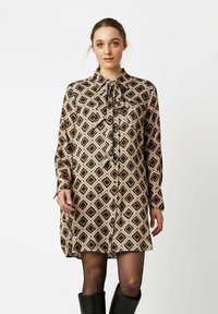 Frau trägt ein langärmliges, knöpfbares Kleid mit einem geometrischen Diamantmuster in Beige, Schwarz und Braun, kombiniert mit schwarzen Strumpfhosen und knielangen Stiefeln.