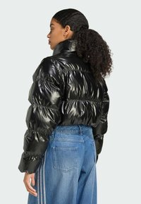 Een jonge vrouw met krullend haar draagt een glanzende zwarte pufferjack en blauwe jeans, gezien van achteren tegen een lichte achtergrond.