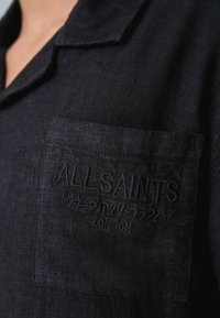 Gros plan de la poche d'une chemise noire avec le texte brodé "ALLSAINTS", des caractères japonais et "LONDON" sur un tissu texturé.