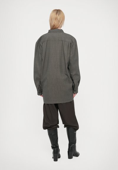 Persona rubia de espaldas, vistiendo una camisa gris oversize, pantalones marrones sueltos metidos dentro de botas de tacón negras hasta la rodilla, sobre un fondo blanco.