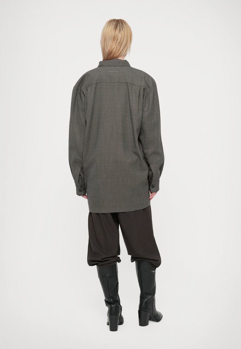 Personne blonde de dos, portant une chemise grise ample, un pantalon marron large rentré dans des bottes noires à talons sur un fond blanc.