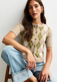 Donna con lunghi capelli castani che indossa un cardigan a maniche corte beige e verde a rombi e jeans chiari arrotolati, seduta contro un muro bianco.