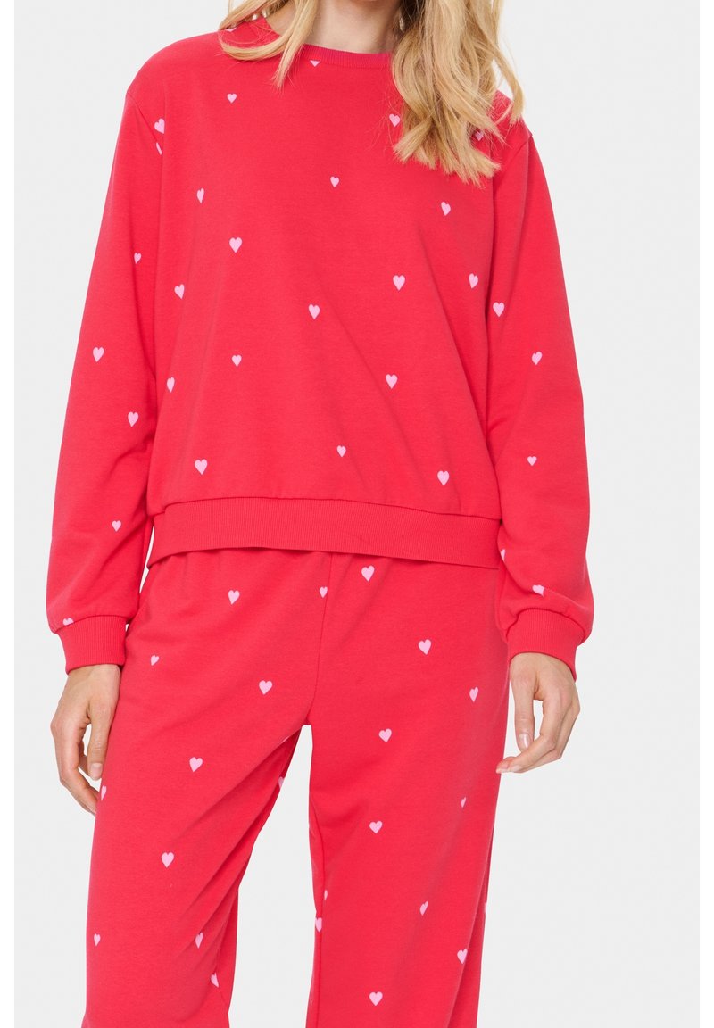 Femme portant un sweat-shirt et un pantalon rouges assortis avec de petits motifs de cœurs blancs sur un fond uni.