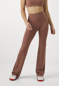 Brune høytliv leggings i et glatt, elastisk stoff, parret med en kort topp. Modellen står med hendene på hoftene, iført sporty sko.