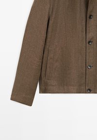 Manche de veste marron à motif chevrons avec fermeture à boutons sur le devant et une poche fendue horizontale en bas.