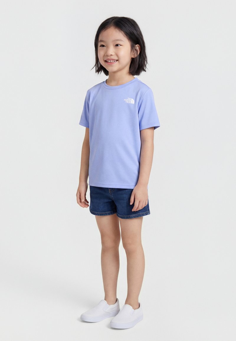 Χαμογελαστό παιδί όρθιο, που φοράει ένα ανοιχτό μοβ κοντομάνικο μπλουζάκι, σκούρα τζιν σορτς και λευκά παπούτσια χωρίς κορδόνια πάνω σε απλό φόντο.