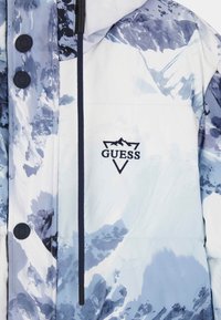 Doudoune bleue et blanche, ornée d'un imprimé montagne. Comprend une capuche, des boutons noirs et un logo GUESS brodé en marine.