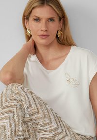 T-shirt blanc à manches courtes avec illustration d'abeille dorée, associé à un pantalon beige et blanc à motif chevron. Texture de tissu lisse.