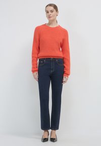 Oranger strukturierten Pullover mit Rundhalsausschnitt, kombiniert mit engen dunkelblauen Jeans. Elegante schwarze spitze Absätze vervollständigen das Outfit.