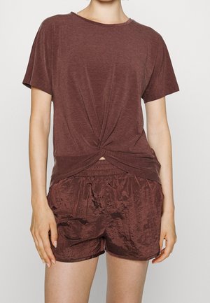 Frau trägt ein kastanienbraunes, kurzärmeliges, geknotetes Top und passende glänzende Shorts vor einem schlichten weißen Hintergrund.