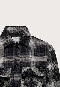 Camisa de franela a cuadros en blanco y negro, con cuello de punta, dos bolsillos en el pecho y cierres de botones. Textura suave con un patrón a cuadros.