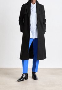 Osoba v dlouhém černém kabátu, modrých kalhotách, černých botách na podpatku a světle modré bílé pruhované košili stojící na neutrální podlaze.