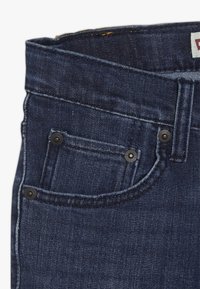 Vaqueros de mezclilla azul oscuro con un diseño clásico, que cuentan con un bolsillo frontal, detalles de botones de metal y costuras visibles.