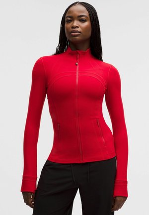 Femeie cu cozi lungi împletite, purtând o jachetă roșie cu fermoar, ajustată, cu găuri pentru degete și pantaloni negri, stând în picioare pe un fundal simplu.