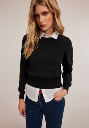 Black knitted sweater with a round neckline, paired over a white button-up shirt. The outfit features blue denim jeans and a bright red belt.