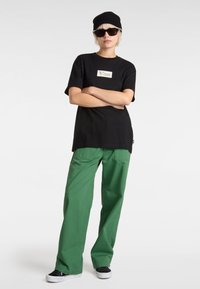 Zwarte katoenen t-shirt met klein Vans-logo, gecombineerd met groene brede broek en zwarte sneakers. Model draagt een zonnebril en een zwarte beanie.
