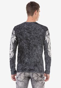 Schwarzes langärmliges Shirt mit strukturierter Oberfläche. Sie verfügt über weiße Schlangengrafiken auf den Ärmeln und filigrane Muster auf dem Rücken.