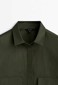 Chemise vert olive avec un col pointu, deux poches poitrine et une texture lisse. Présente un devant à boutonnière et des manches courtes.