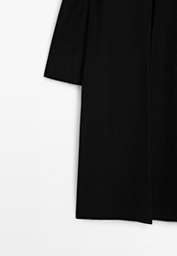 Schwarzer langer Mantel mit herunterhängendem Schulternaht, sichtbarer vertikaler Naht vorne und geradem Saum auf weißem Hintergrund.