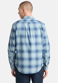 Camicia a maniche lunghe di colore azzurro chiaro con un motivo a quadri in blu scuro. Caratterizzata da un dietro con spacco e polsini con bottone. Tessuto in cotone con texture liscia.