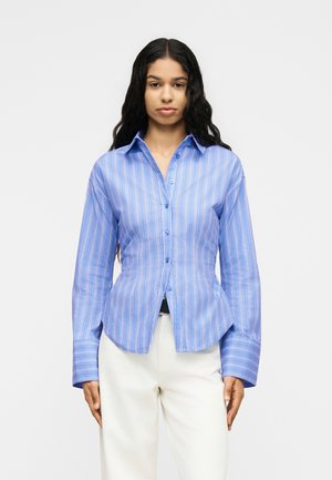 Nő, hosszú göndör hajjal, kék csíkos, gombos ingben és fehér nadrágban, egyszerű világos háttér előtt áll.