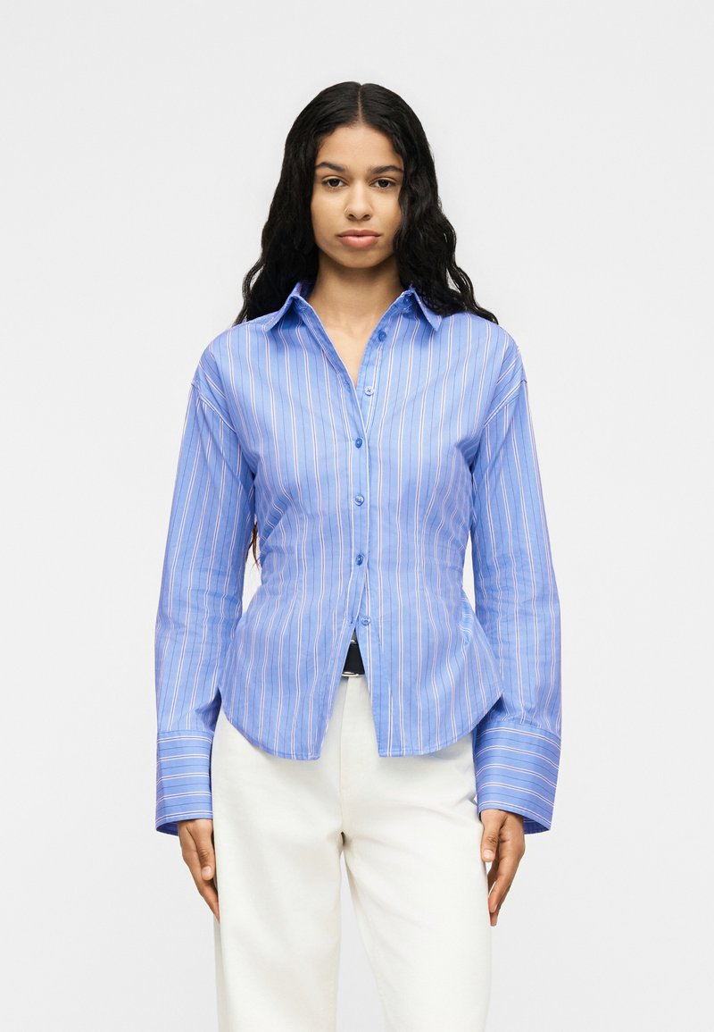 Γυναίκα με μακριά σγουρά μαλλιά που φοράει μπλε ριγωτό πουκάμισο με κουμπιά και άσπρο παντελόνι, στέκεται μπροστά σε ένα απλό φωτεινό φόντο.