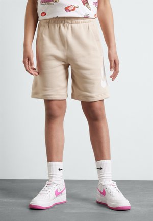 Dziecko w beżowych szortach z białym logo Nike, białych skarpetkach i białych butach sportowych z różowymi logo Nike, stojące na szarej podłodze.