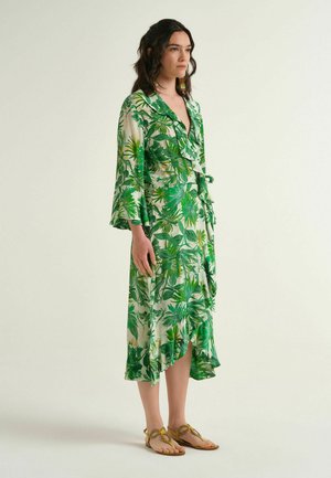 Woman wearing a midi wrap dress with green tropical leaf print, flared sleeves, and beige sandals, standing against a plain light background.