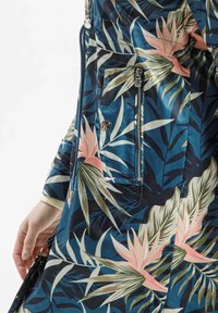 Kurtka w tropikalny wzór na niebieskim tle, z różowymi i zielonymi wzorami liści, z kieszeniami na zamek błyskawiczny oraz gładką, błyszczącą fakturą.