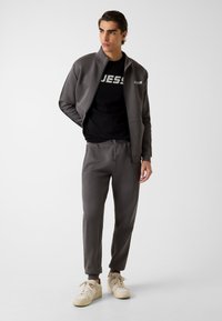 Giacca grigia con cerniera e dettagli neri, abbinata a una t-shirt nera con la scritta "GUESS" e pantaloni jogging grigi coordinati; sneakers color crema.