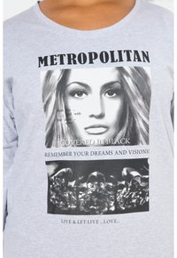 Grijze langarmige t-shirt met een bedrukte afbeelding van een vrouwen gezicht, tekstoverlays en een afbeelding van zwarte elementen eronder.