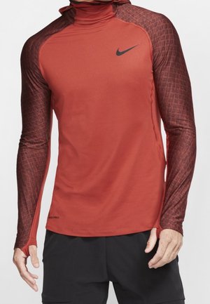 Mand iført rustfarvet Nike langærmet træningstop med strukturerede ærmer og sort swoosh-logo på brystet, kombineret med sorte shorts.