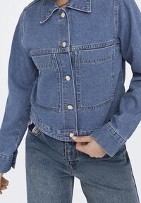 Jeansjacka i ljusblått med knappslutning fram, två bröstfickor och en spetsig krage. Distinkt sömnad längs sömmarna och manschetterna.