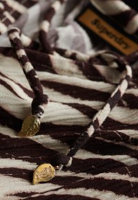 Tejido a rayas marrón y crema con una sensación texturizada y ligera. Presenta un cordón con acentos en forma de hoja dorada y una etiqueta de logo.