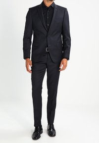 Traje oscuro hecho a medida de tela lisa, que incluye un saco de un solo botón, pantalones a juego y zapatos de cuero negros.