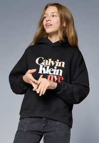Čierna mikina s kapucňou s predným grafickým motívom "Calvin Klein nyc" v béžovej a červenej farbe. Má sťahovaciu kapucňu a rebrované manžety.