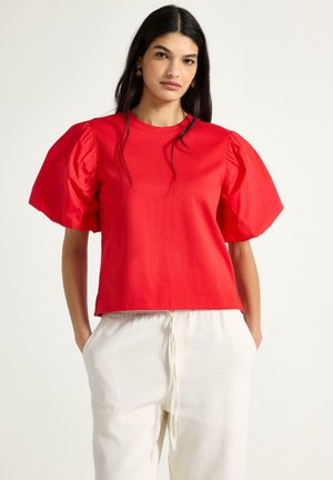 Kvinde med langt mørkt hår iført en rød bluse med pufærmer og hvide bukser med snøre, stående med hænderne i lommerne foran en ensfarvet baggrund.