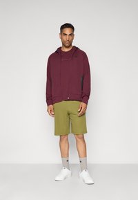 Mann in einer weinroten Zip-Hoodie, passendem weinroten T-Shirt, olivgrünen Shorts, grauen Logostrümpfen und weißen Sneakers, der vor einem schlichten Hintergrund steht.